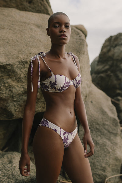 Woman wearing a palm print bikini set in purple and white standing against rocky terrain