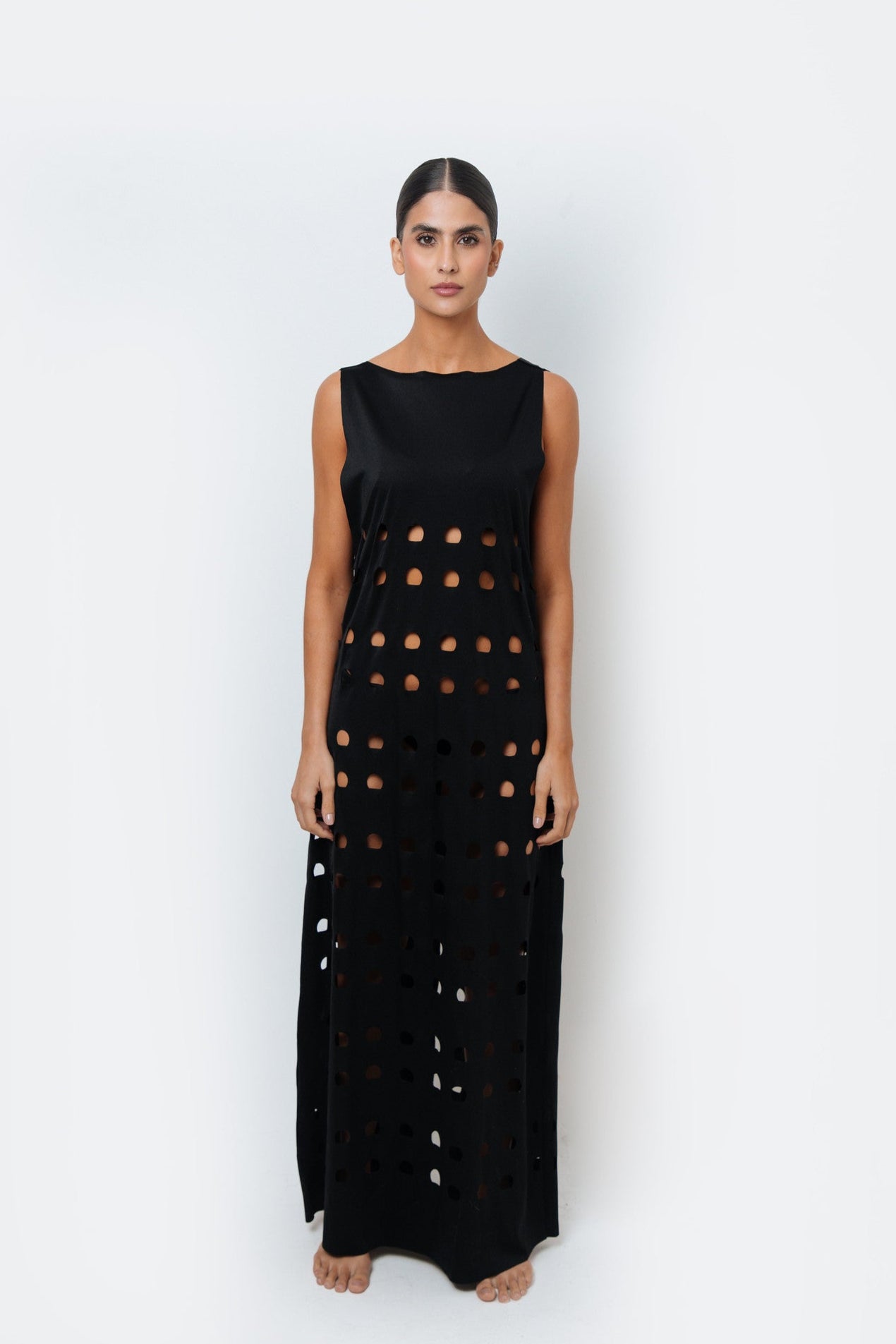 Model wearing a sleeveless black maxi dress with circular laser-cut detailing from waist to hem, part of WOAHCO’s statement resortwear collection.